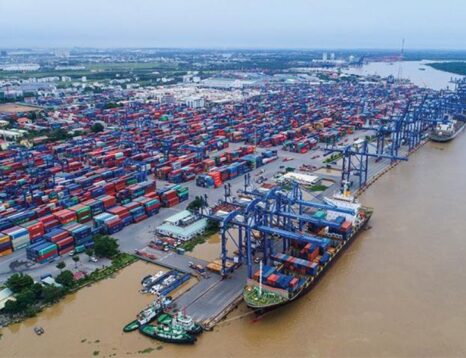 Aerial view of a busy shipping port with stacked colorful containers, blue gantry cranes, and cargo ships along a brown river.