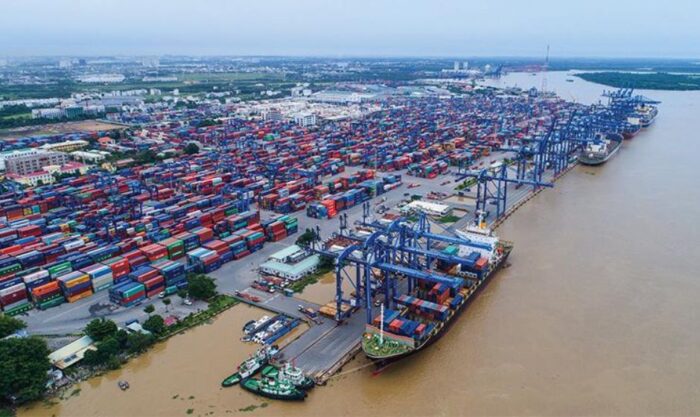 Aerial view of a busy shipping port with stacked colorful containers, blue gantry cranes, and cargo ships along a brown river.