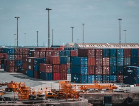 Stacks of multicolored shipping containers at a dockyard with orange ground equipment in the foreground.