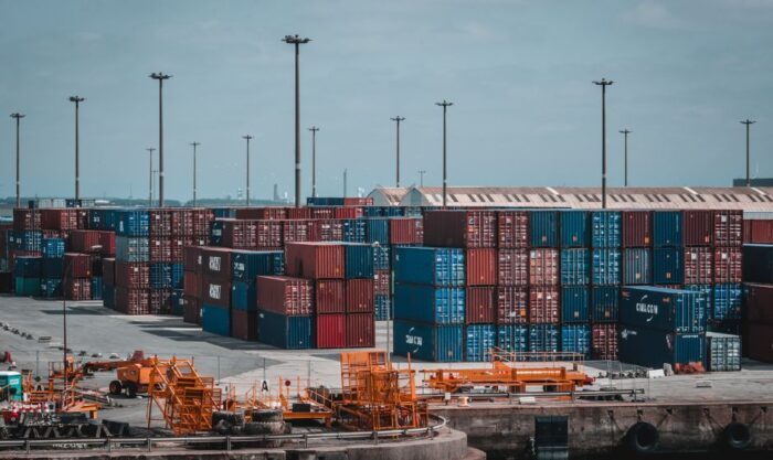 Stacks of multicolored shipping containers at a dockyard with orange ground equipment in the foreground.