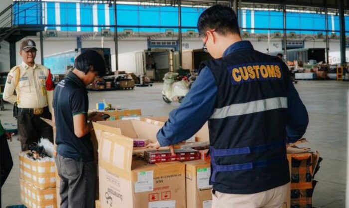 Customs officer in a vest inspects boxes on a table inside a large warehouse; other workers oversee the process.