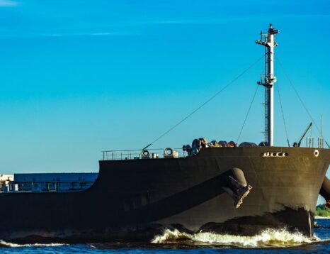 Large dark cargo ship sailing in blue water under a clear blue sky, with a tall mast and deck equipment visible at the bow and stern area.