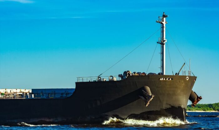 Large dark cargo ship sailing in blue water under a clear blue sky, with a tall mast and deck equipment visible at the bow and stern area.
