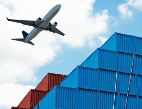Airplane flying over stacked blue and red shipping containers in a port-like yard.