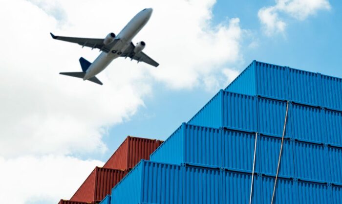 Airplane flying over stacked blue and red shipping containers in a port-like yard.