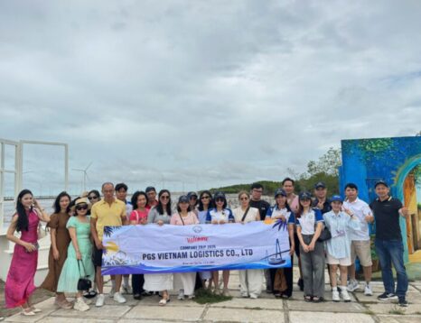 Group of employees posing outdoors on a waterfront, holding a banner reading 'PGS VIETNAM LOGISTICS CO., LTD' during a company trip.
