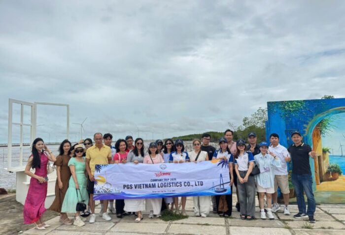 Group of employees posing outdoors on a waterfront, holding a banner reading 'PGS VIETNAM LOGISTICS CO., LTD' during a company trip.