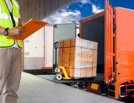 Warehouse worker in a high-visibility vest with clipboard supervising a pallet being loaded onto an orange truck at a loading dock.