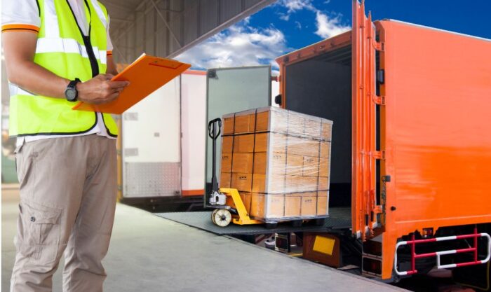 Warehouse worker in a high-visibility vest with clipboard supervising a pallet being loaded onto an orange truck at a loading dock.