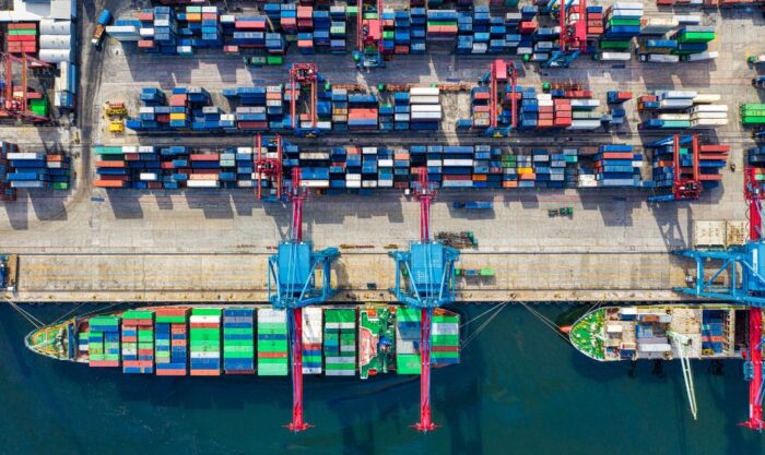 Aerial view of a busy shipping port with stacked colorful containers and large blue gantry cranes over the docked ships left and right