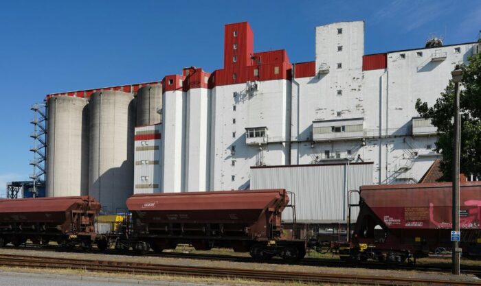 Grain silo complex with red and white buildings behind a line of freight train cars on tracks under a blue sky